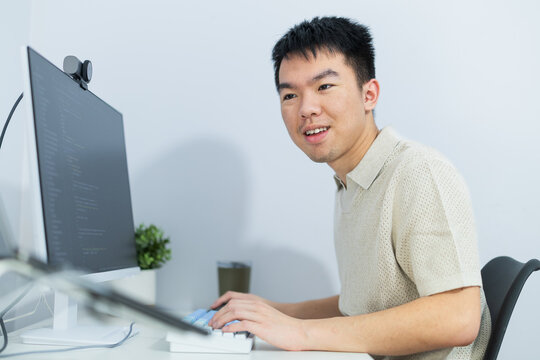 A young Asian man codes at a home workstation, for software development, technology careers, remote employment, or digital skills training.