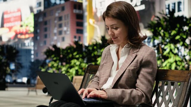 Smiling businesswoman finishes work tasks in a city park and closes her laptop, pleased after finalizing a project while sitting on a wooden bench.