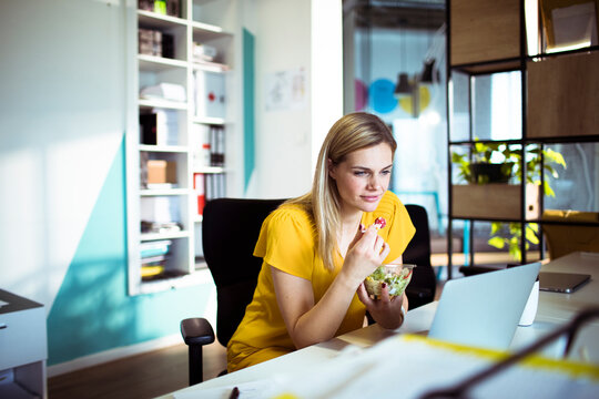 Young woman eating healthy lunch while working on laptop in modern office
