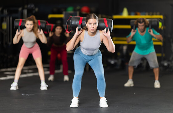 Determined young woman performing sandbag squats during group crossfit workout in gym