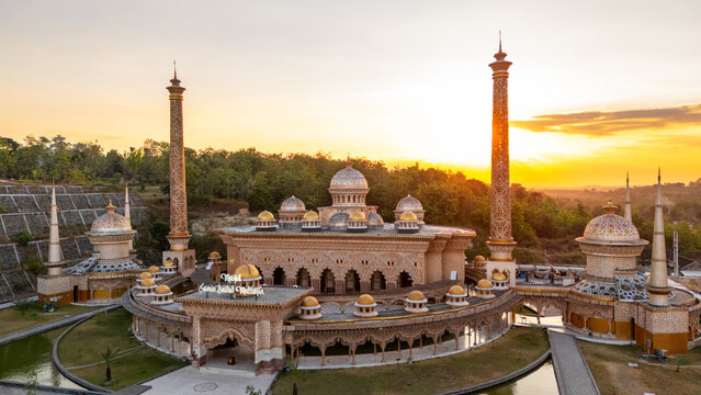 Aerial view of the ornate Samin Baitul Muttaqin Mosque with its golden domes and tall minarets, surrounded by lush trees and a reflecting pond at sunset in Bojonegoro, East Java, Indonesia.