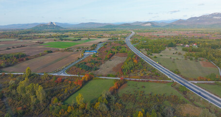 Naklejka premium AERIAL: Motorway interchange surrounded by colourful autumn landscape in Croatia. Roads, fields, scattered woodland and distant hills merge into a scenic blend of nature and modern infrastructure.