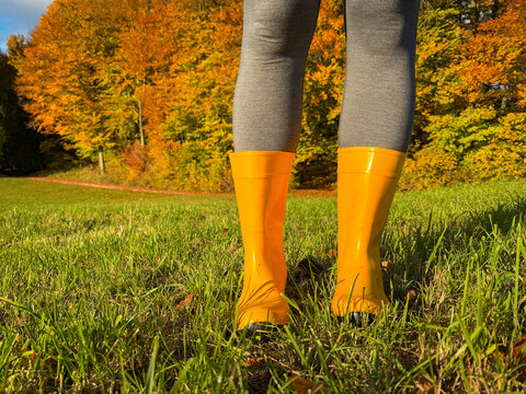 LOW ANGLE VIEW, CLOSE UP: Woman in yellow rain boots stands on green grass beside glowing autumn forest. Seasonal walk through countryside lit by warm sunlight and coloured in vibrant fall foliage.