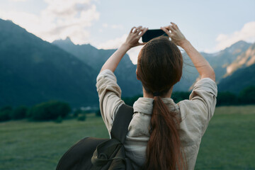 Naklejka premium A woman outdoors with a backpack photographs distant mountains, capturing a moment of exploration and freedom. A calm lifestyle scene emphasizing nature, travel, and personal discovery.