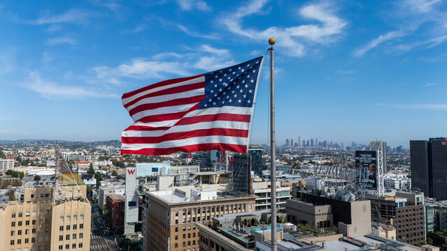 Aerial view of the American flag waving in the wind above the urban skyline of Hollywood with buildings and city streets under a clear blue sky in Los Angeles, California, United States.