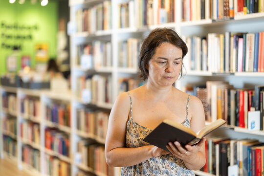 Focused middle-aged lady browsing through literature volumes on shelves in cozy bookshop