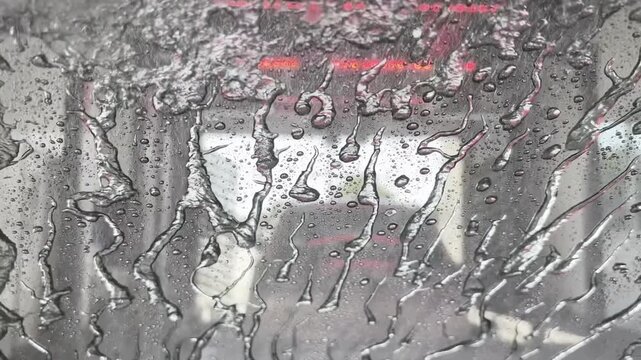 A forceful final rinse sprays across a vehicle windshield beneath red tunnel lights near the exit of an automatic car wash.