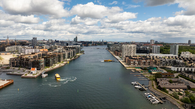 Aerial view of the modern waterfront with yellow water buses navigating the canal, surrounded by contemporary apartment buildings under a cloudy sky in Copenhagen, Denmark.