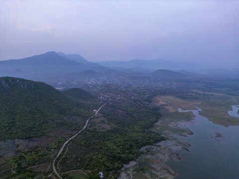 Aerial view of a landscape featuring a winding road, lush green hills, and a lake under a soft morning sky in Jalapa, Oaxaca, Mexico.
