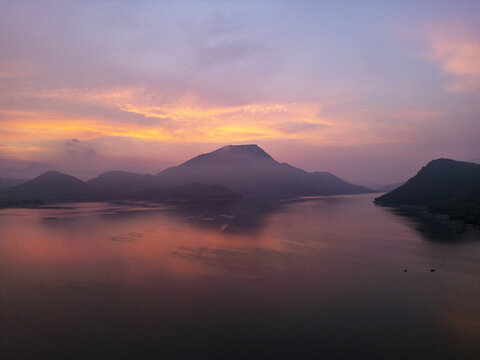 Aerial view of a serene lake at sunrise with mist-covered mountains reflecting in the calm water under a vibrant orange and purple sky in Jalapa, Oaxaca, Mexico.