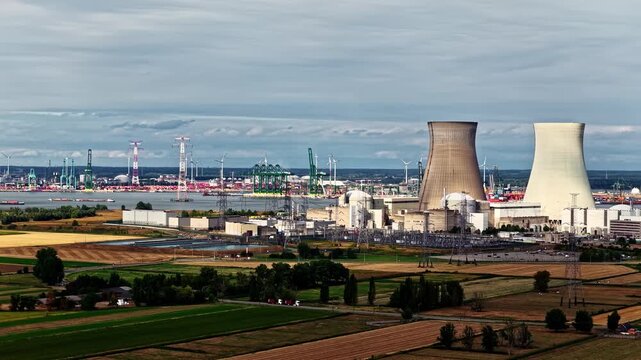 Doel Nuclear Power Station cooling towers rise above farmland with the busy Port of Antwerp cranes in the distance under cloudy skies.
