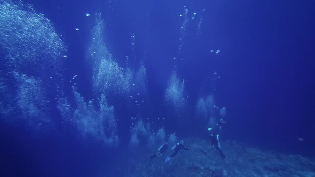 Group of scuba divers swims along the seafloor as large streams of air bubbles rise dramatically through deep blue water.