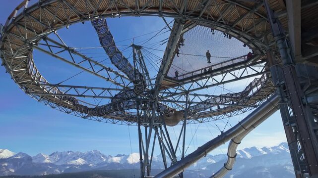 SkyWalk Serce Poronina in Male Ciche, Poland, with families on the elevated lookout walkway, slide and net bridge above snowy Tatra mountain scenery on a sunny winter day.