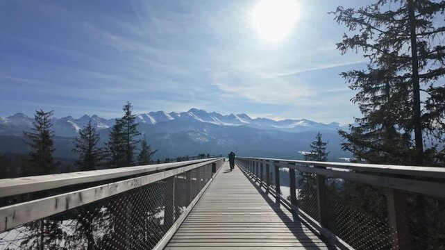 SkyWalk Serce Poronina in Male Ciche, Poland, with tourists on a wooden elevated walkway above evergreen forest and views of the snow-capped Tatra Mountains on a sunny winter day.