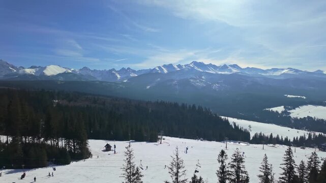 SkyWalk Serce Poronina in Male Ciche, Poland, with ski lifts and skiers on snowy slopes beneath the Tatra Mountains, surrounded by coniferous forest under a clear blue sky.
