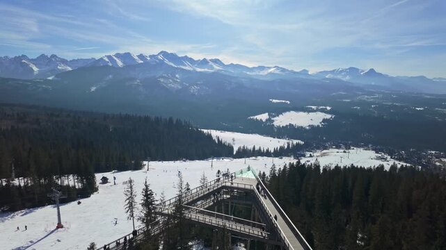 SkyWalk Serce Poronina in Male Ciche, Poland, with wooden treetop walk and observation deck overlooking snowy Tatra Mountains peaks, ski slopes, forest, and winter landscape.