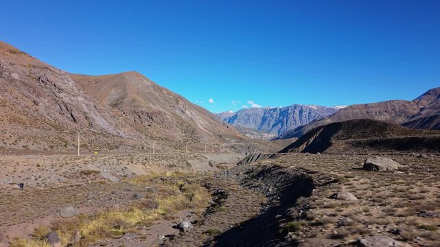Low lateral drift along a dusty mountain valley road bordered by dry rocky slopes and scattered scrub with distant snow capped Andean peaks rising on the clear far horizon