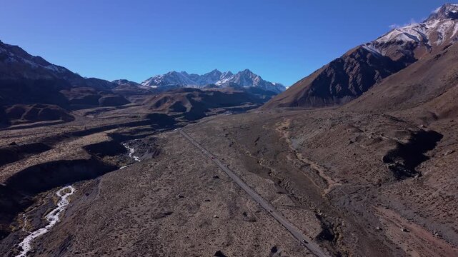 Backward retreating flight revealing a straight mountain highway cutting through a barren plateau beside a winding river toward sharp jagged snow capped Andean peaks