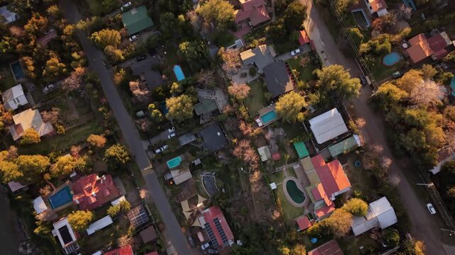 Top down nadir shot of a suburban residential neighborhood with colorful rooftops, backyard swimming pools, and golden autumn trees lining quiet streets at sunset
