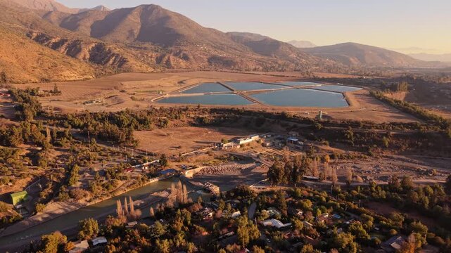 Forward flight over a small mountain town and winding tree lined river toward distant geometric water treatment ponds framed by golden foothills at warm sunset