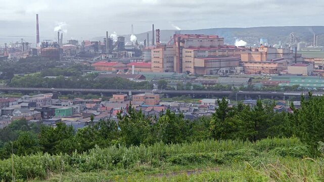 丘の上から煙を上げる工場を望む風景。