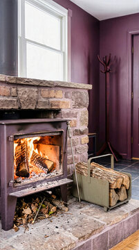 Burning wood logs inside cast iron stove in brick fireplace hearth. Vertical interior view of home heating with metal firewood basket, iron poker tool, kindling and purple wall background