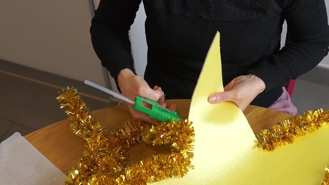 Close-up video of a woman adding hot glue to a fluffy golden tinsel cluster during a DIY handcrafting session. Perfect concept for Christmas ornaments, party props, or carnival parade preparation.