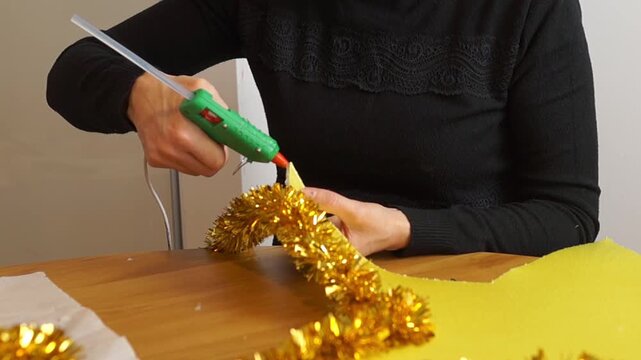 Detailed shot of female hands carefully pressing and fixing golden shiny tinsel onto a large yellow cutout star using thermal adhesive.