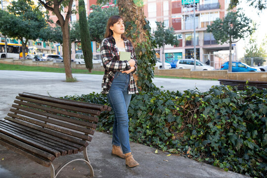 young woman walking through city park with a book