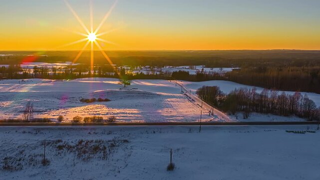 Low flyover along a country road as the sun sets over patchy snow and bare trees across open farmland.