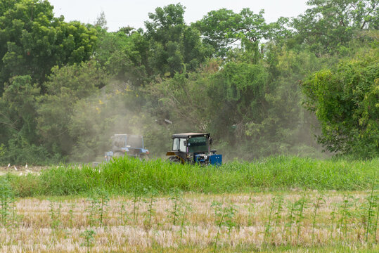 Two farm tractors working across rural field surrounded by green vegetation trees dust rising behind agricultural machinery during land cultivation