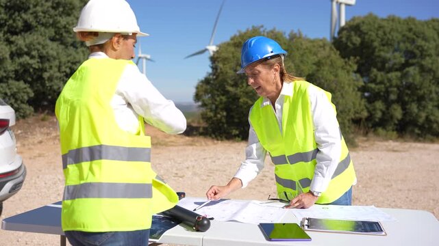 Two female engineers in hard hats and safety vests discussing blueprints at a wind turbine farm