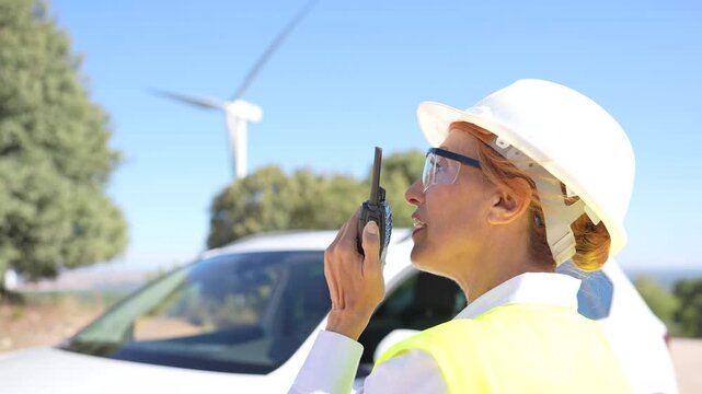 Woman in a hard hat using a walkie talkie at a renewable energy site with a wind turbine