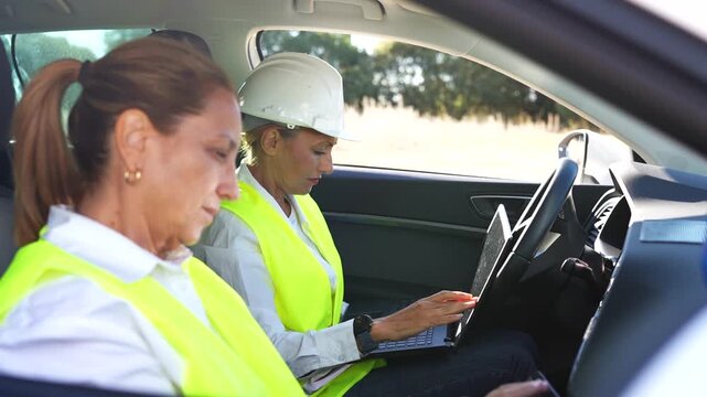 Two women engineers in safety vests and hard hats working on a laptop inside a car on a sunny day
