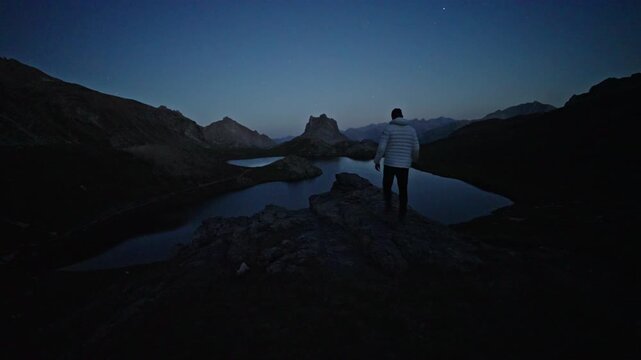 A lone hiker in a white jacket stands on a rocky ridge, silhouetted against twin mountain lakes at blue hour.