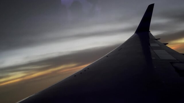 Airplane wing silhouette flying through beautiful clouds at sunset with a dramatic sky view from the passenger window of the commercial aircraft during the flight