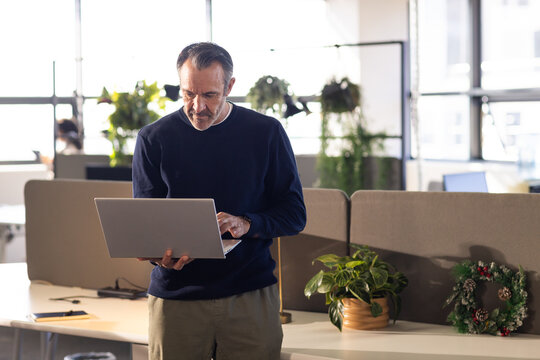 Middle-aged man standing by fabric partition in open-plan office, typing on laptop with wreath