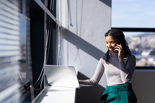 African American woman standing in office by blinds holding phone to ear, touching laptop on ledge
