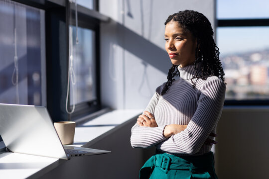 African American woman standing at office window ledge with silver laptop, ceramic mug, copy space