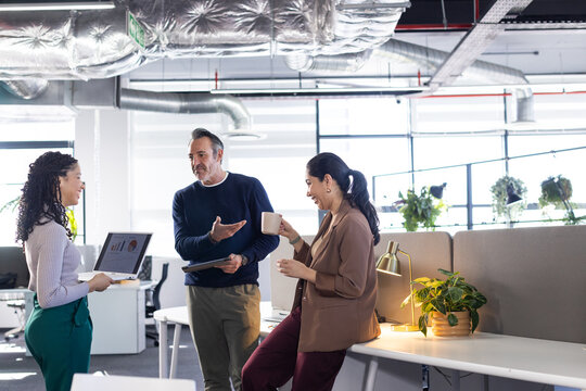 Diverse coworkers collaborating over data charts at office desk with laptop, tablet, coffee mug