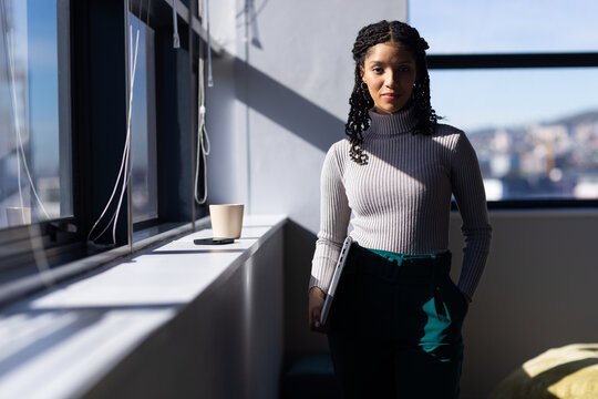 African American woman in sweater standing by windowsill holding laptop with coffee cup, copy space
