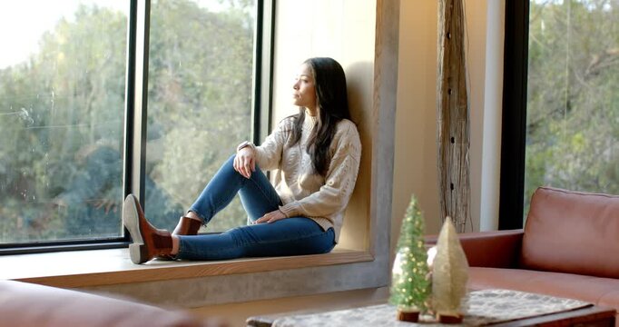 Woman sitting on living room window ledge gazing at trees beyond beside tabletop trees, copy space