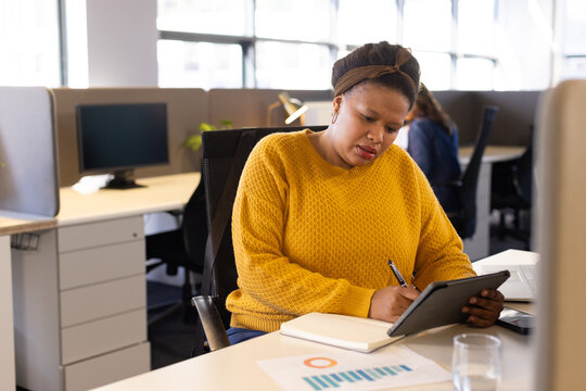 Diverse coworkers writing on digital tablet with stylus near notebook and chart in open plan office