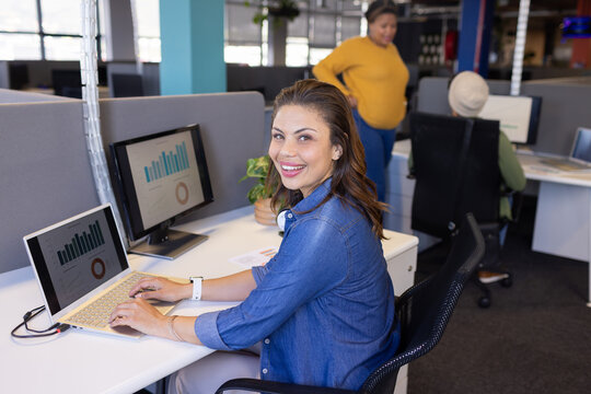 Woman smiling and typing on laptop at modern open plan office with monitors showing charts