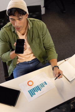 Asian male office worker speaking into smartphone and taking notes on printed charts at desk