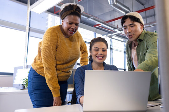 Diverse coworkers collaborating around silver laptop at desk in open office with overhead lights