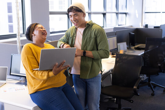 Diverse coworkers laughing and pointing at silver laptop on white desk in open-plan office
