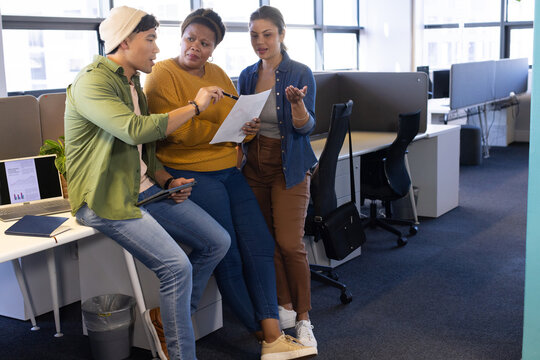 Diverse coworkers collaborating at desk in open office reviewing printed sheet with tablet and pen