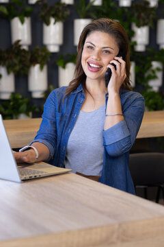 Woman smiling while holding smartphone and typing on laptop at coworking space with white planters