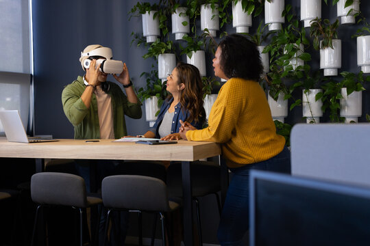 Diverse coworkers adjusting virtual reality headset, leaning on table in office with laptop, plants